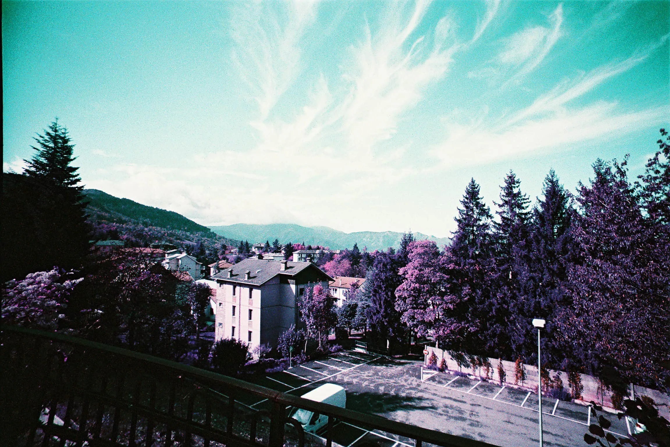 Lomo Purple film photograph overlooking a mountain village, with purple trees, dark rooftops, hills, and a bright blue sky.