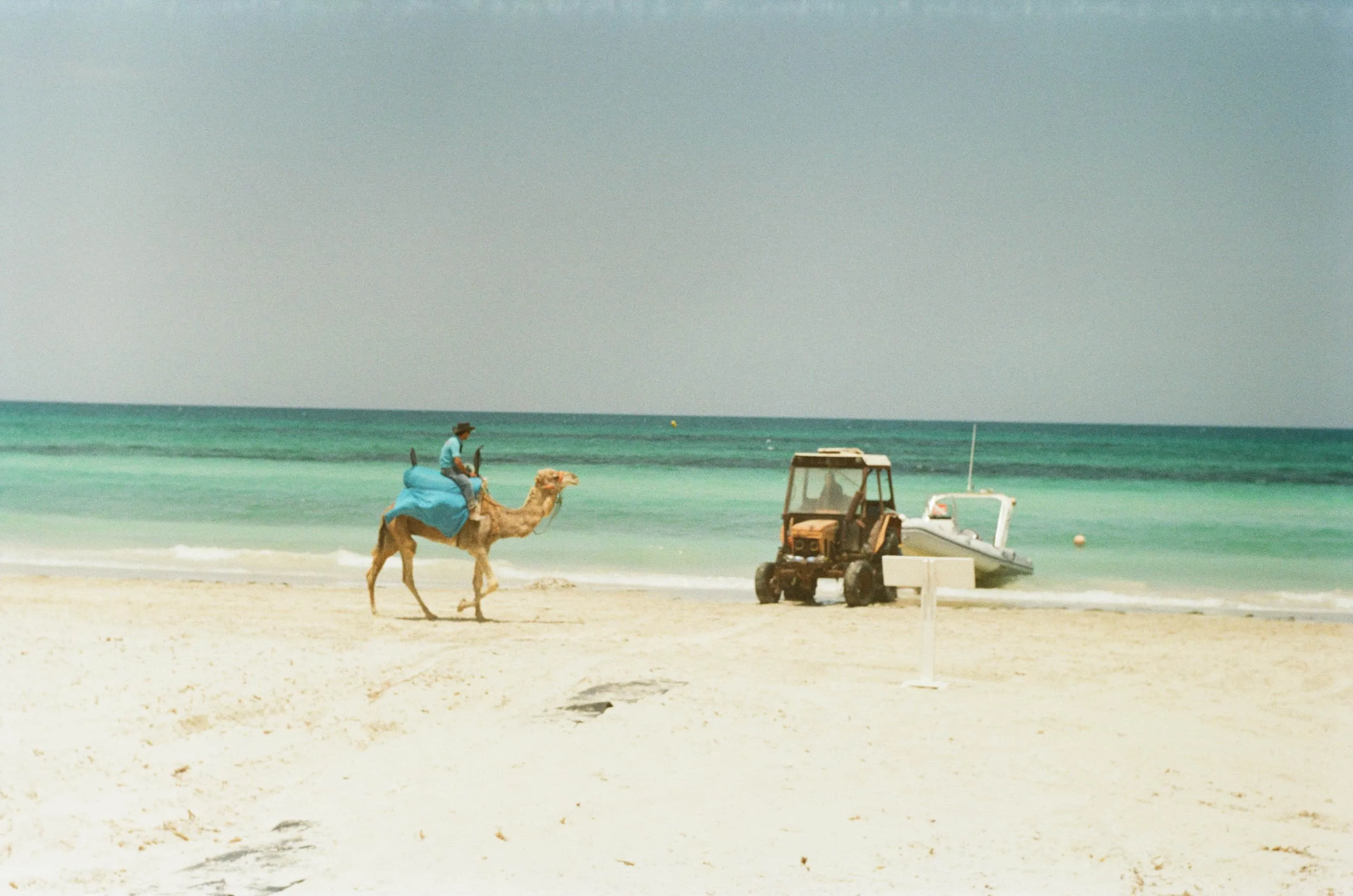 Soft colour film photograph of a camel rider on a beach in Djerba, with turquoise water, a tractor, and a small boat nearby.
