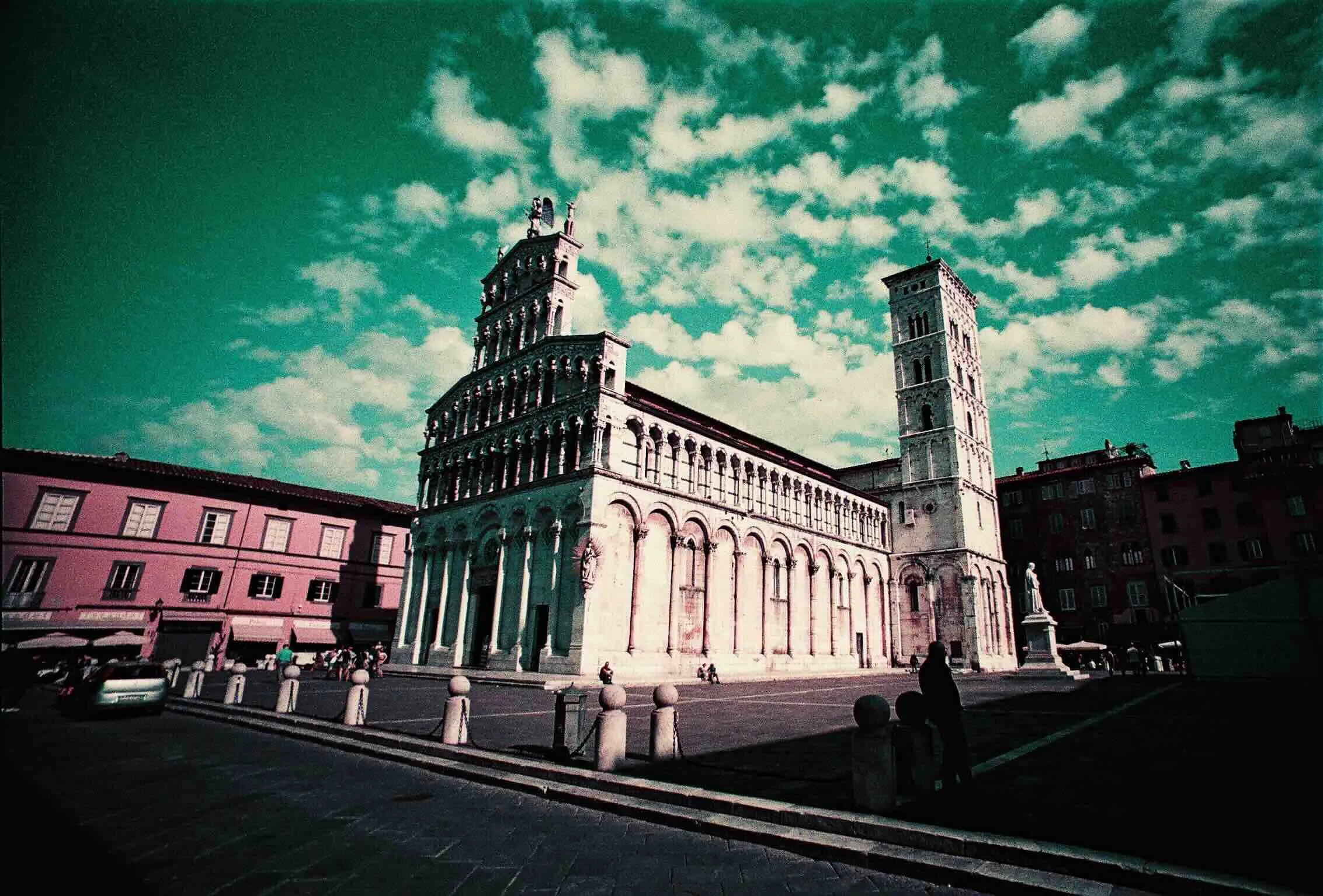 Lomo Purple film photograph of an Italian church and bell tower in a sunlit square under a green-blue sky.